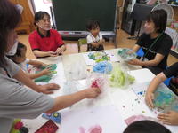 テーブルを囲む幼児と教員。好きな色の紙粘土を手に取り、厚紙の上にのせている。
