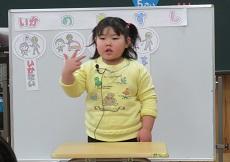 幼児が「し」の指文字（親指、人差し指、中指を出す）を表す。ホワイトボードには、「いかのおすし」の文字カードとそれぞれの音から始まる言葉の絵カードが貼られている。