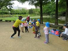 公園の池の前で、横一列に並んだ子どもたち。前に立つ教員が一人の子どもに手を伸ばしている。