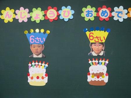 誕生会の壁面。花型の画用紙に描かれた「うじょうびおめで」の文字。６本のろうそくを立てたケーキ２つ。その上に男の子と女の子の顔写真。それぞれの頭の上には、６さいと書かれた冠。