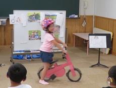 ホワイトボードに自転車の練習をしている時の写真。幼児が桃色のヘルメットをかぶり、手作りの桃色の自転車にまたがる。