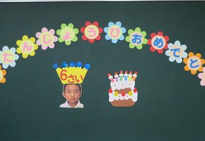 黒板。花形の画用紙に書かれた、おたんじょうびおめでとうの文字、６本のろうそくが立ったペープサートのケーキ。男の子の顔写真の上には６さいと描かれた冠。