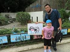 ペンギンの檻の前で、子どもが描いたペンギンの絵を持つお父さんと幼児。