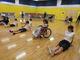 体育館で子供たちが床に座って体操をしている。