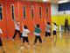 体育館で子供たちがダンスインストラクターの動きをまねて片手を上げてダンスを踊っている。