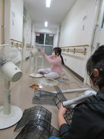 女子２人が廊下で扇風機とエアコンフィルターを掃除しているところ